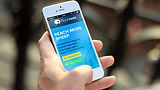 A hand holds a smartphone displaying a blue screen with a small green button, against a blurred outdoor background.